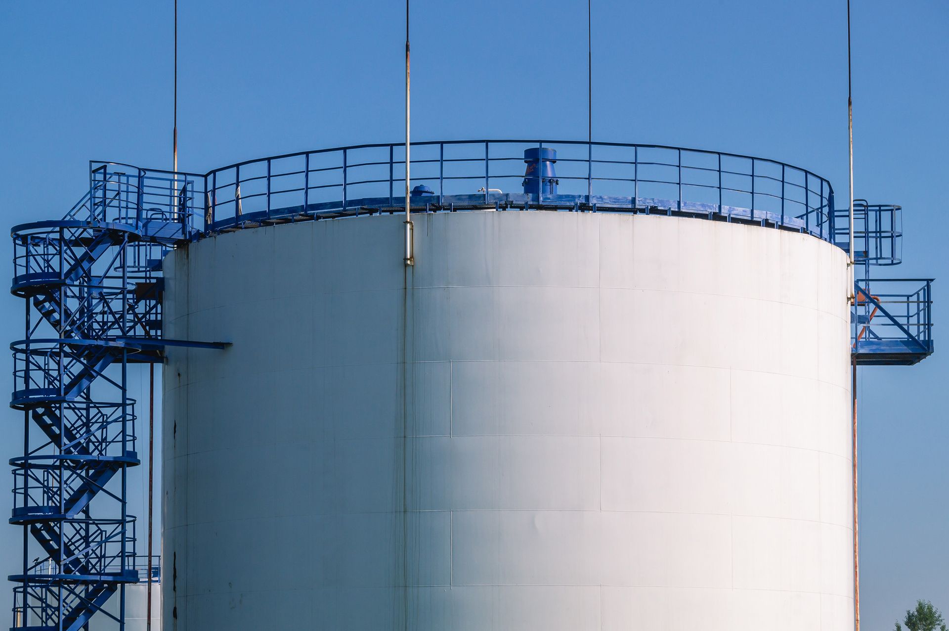 Ein großer, weißer, zylindrischer Industrietank mit blauer Metalltreppe, Blitzableitern und Sicherheitsgeländer vor klarem, blauem Himmel.