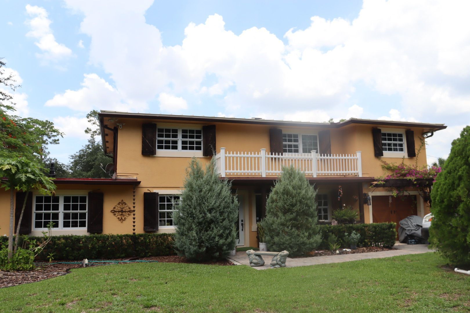 Two-story yellow stucco house with white shutters and a small balcony, trees in front, on a sunny day.
