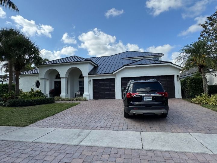 White stucco house with black roof and doors, SUV parked in brick driveway.