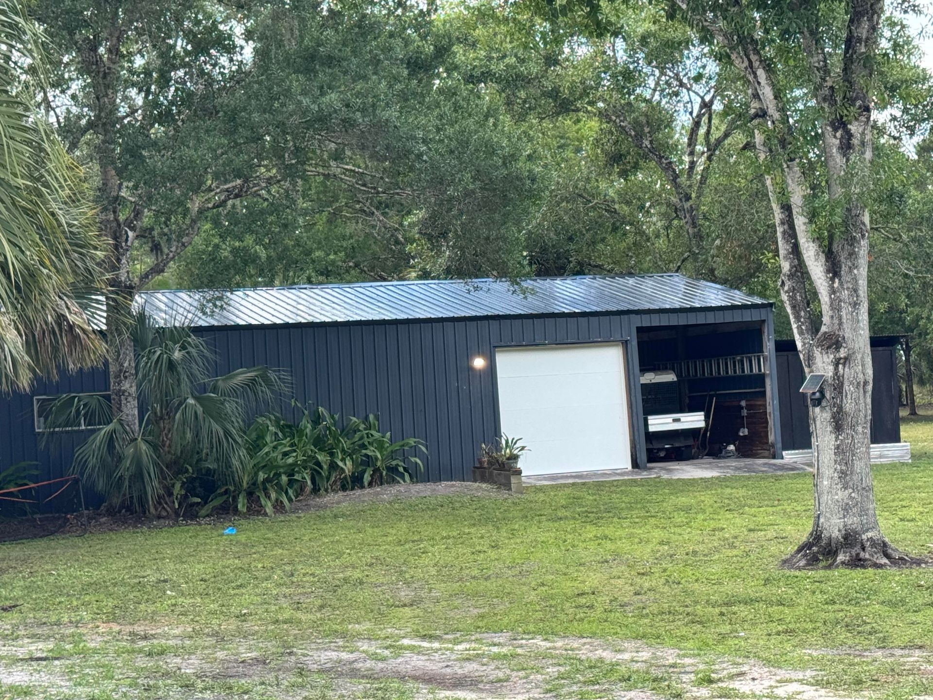 Black shed with white garage door, metal roof, parked vehicle inside, set in a grassy yard, trees in the background.