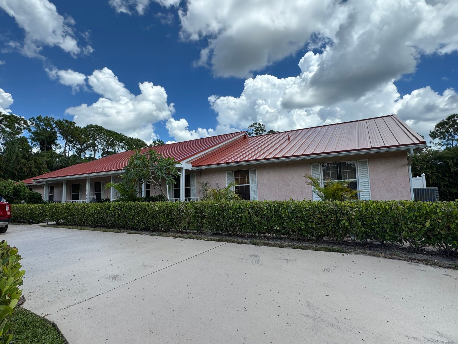 Pink house with red roof under a cloudy blue sky. Hedge lines the driveway.