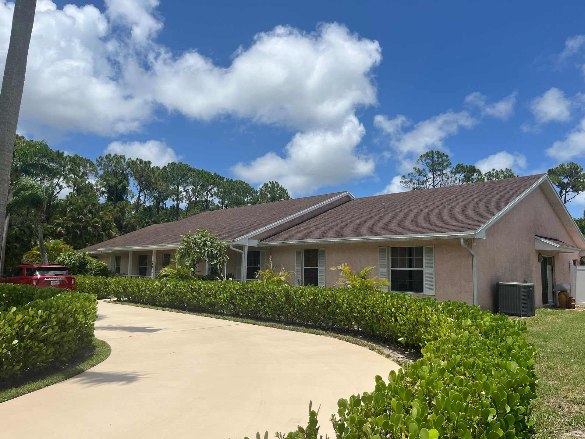 A long, light pink house with a brown roof and a curved driveway surrounded by green bushes under a blue sky.