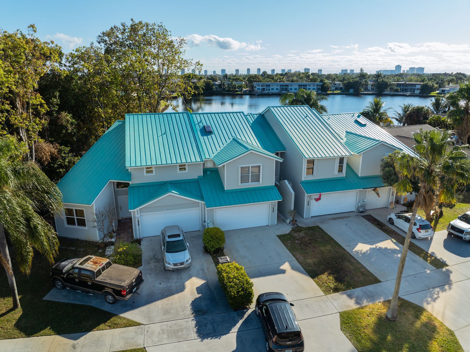 Houses with teal roofs and garages, parked cars, near a lake on a sunny day.