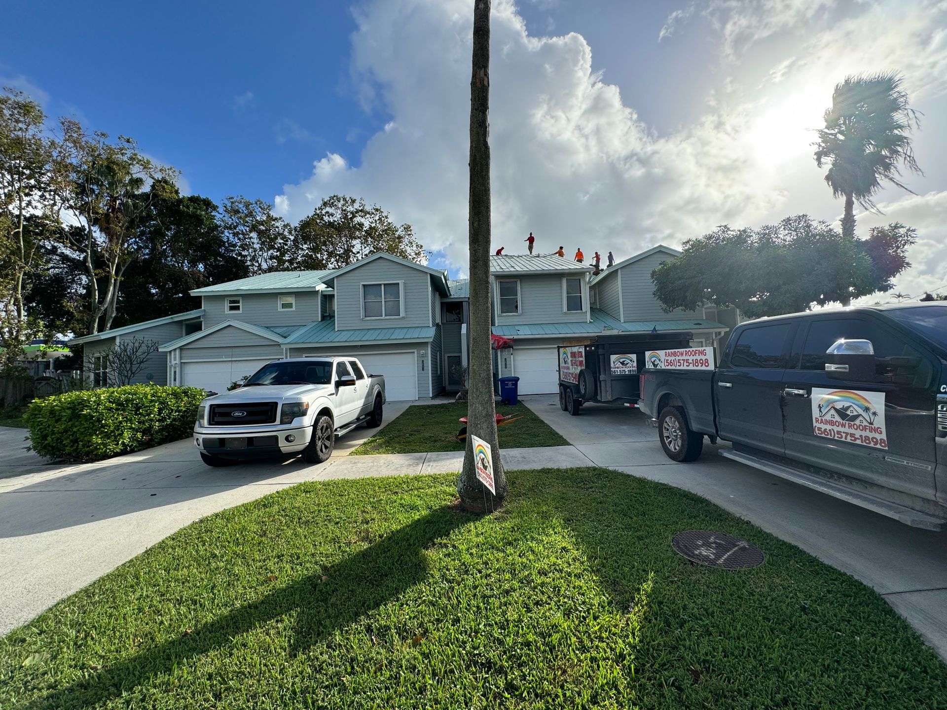 Two-story townhouses with a white truck, black truck, and workers on the roof under a sunny sky.