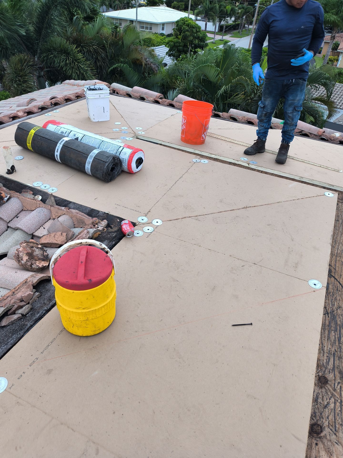 Man on a roof, preparing roofing underlayment. Buckets, rolls, and fasteners are present.