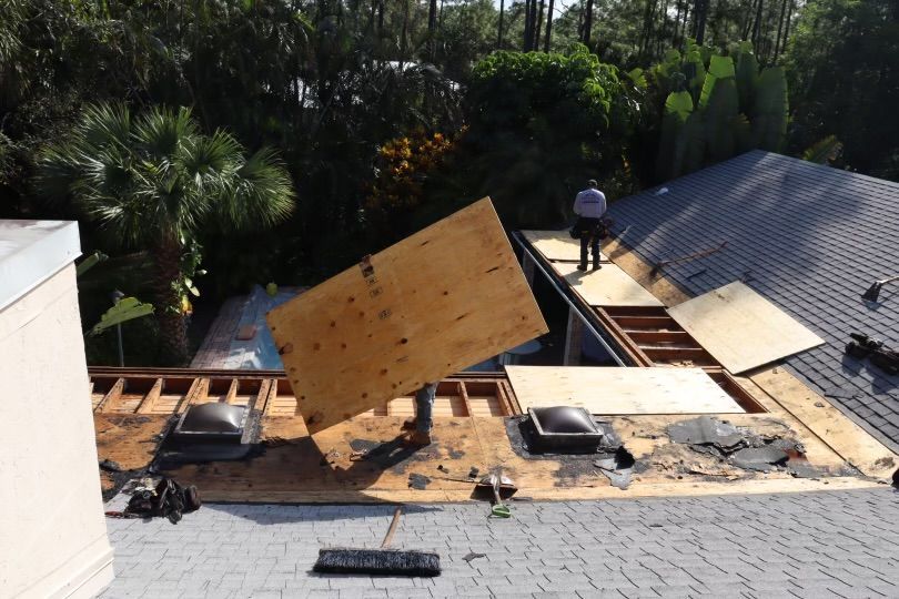 Construction worker lifting plywood on a roof with exposed beams and skylights; sunny day.