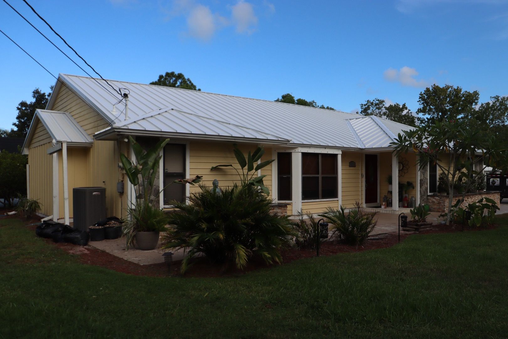 Yellow house with white metal roof, plants, and blue sky.
