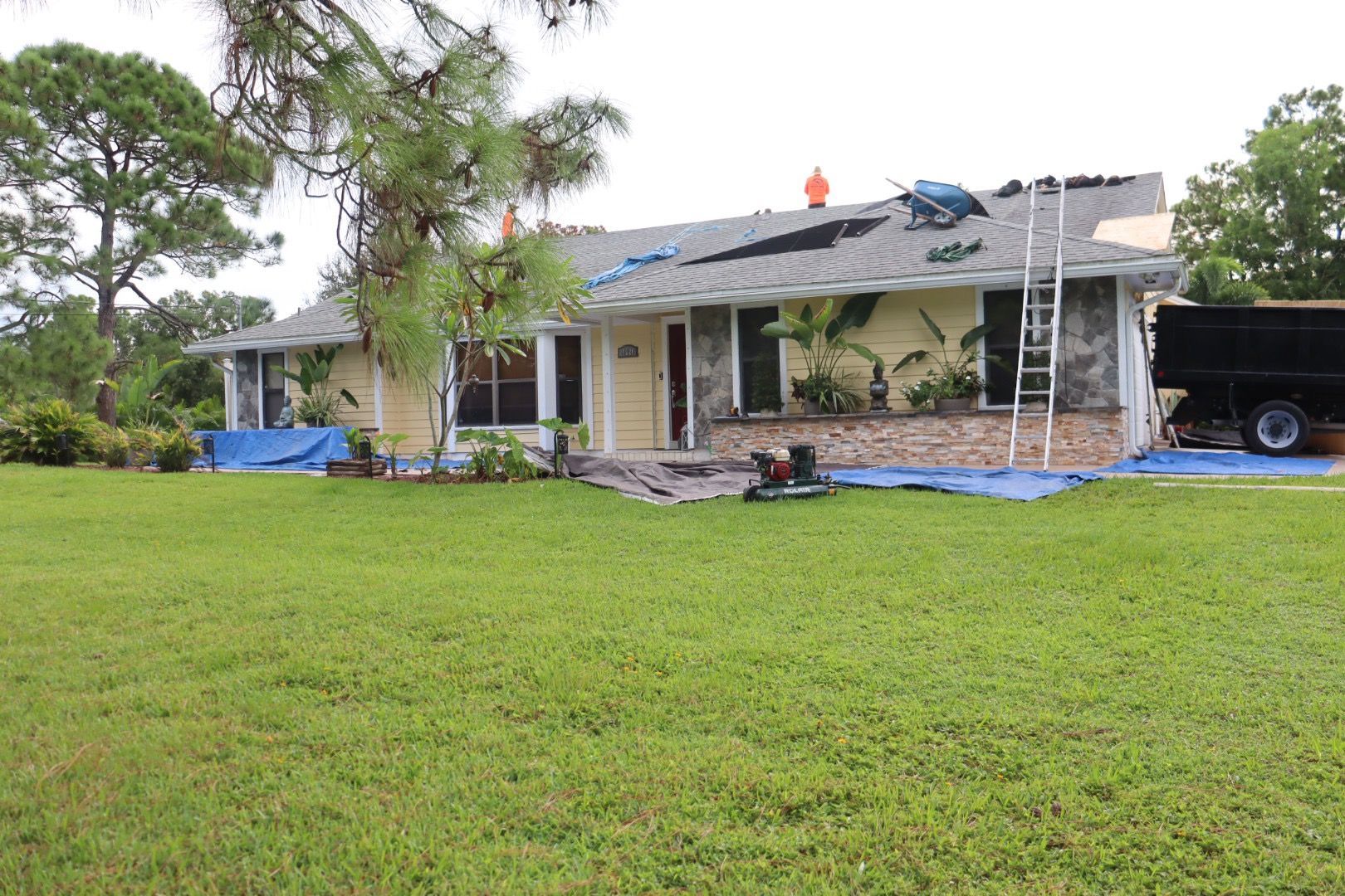 House with roof under repair; blue tarp covers ground; ladder, truck, and workers visible.
