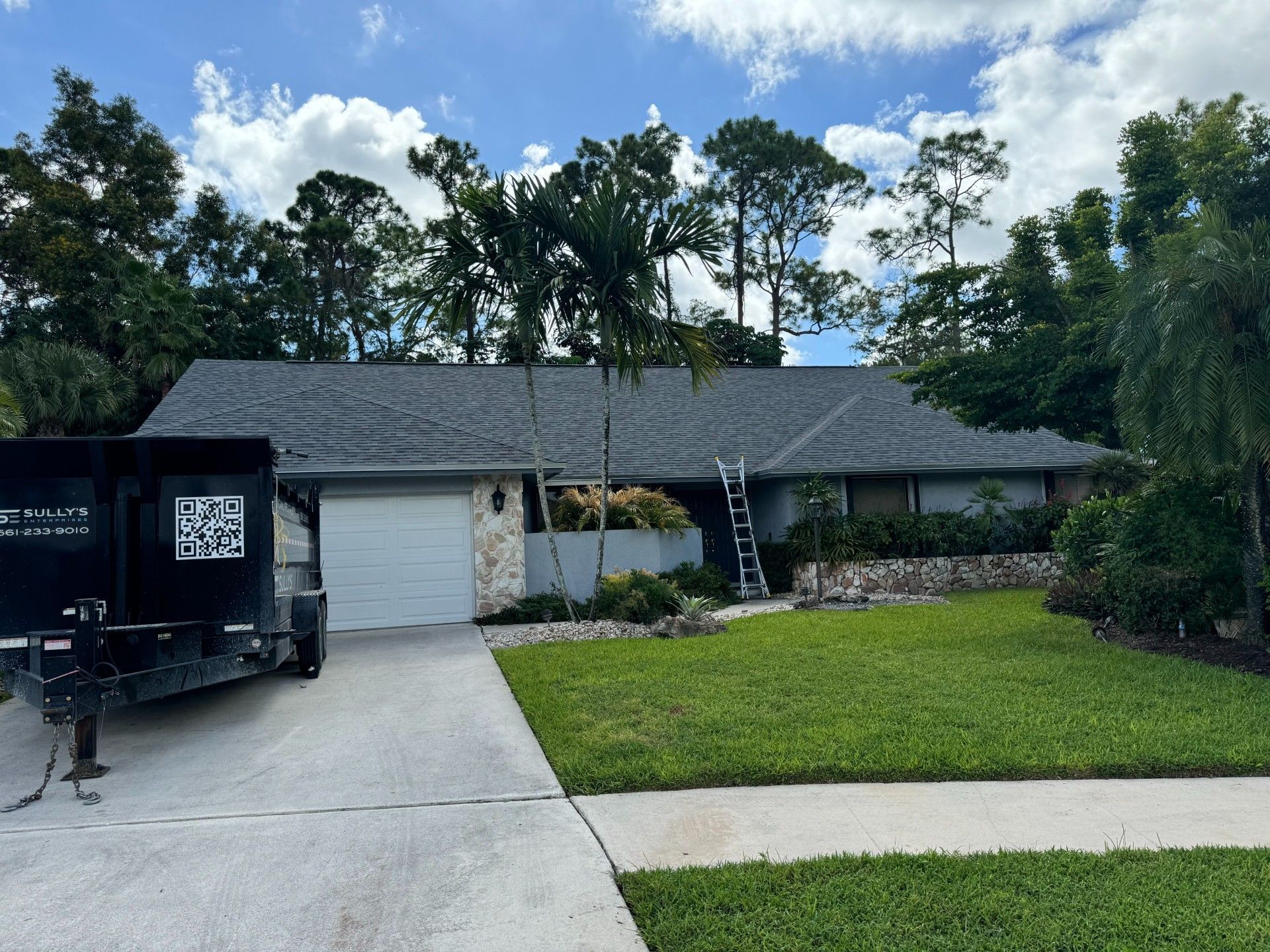 A house with a gray roof, a white garage door, and a trailer in the driveway. Green lawn and trees.