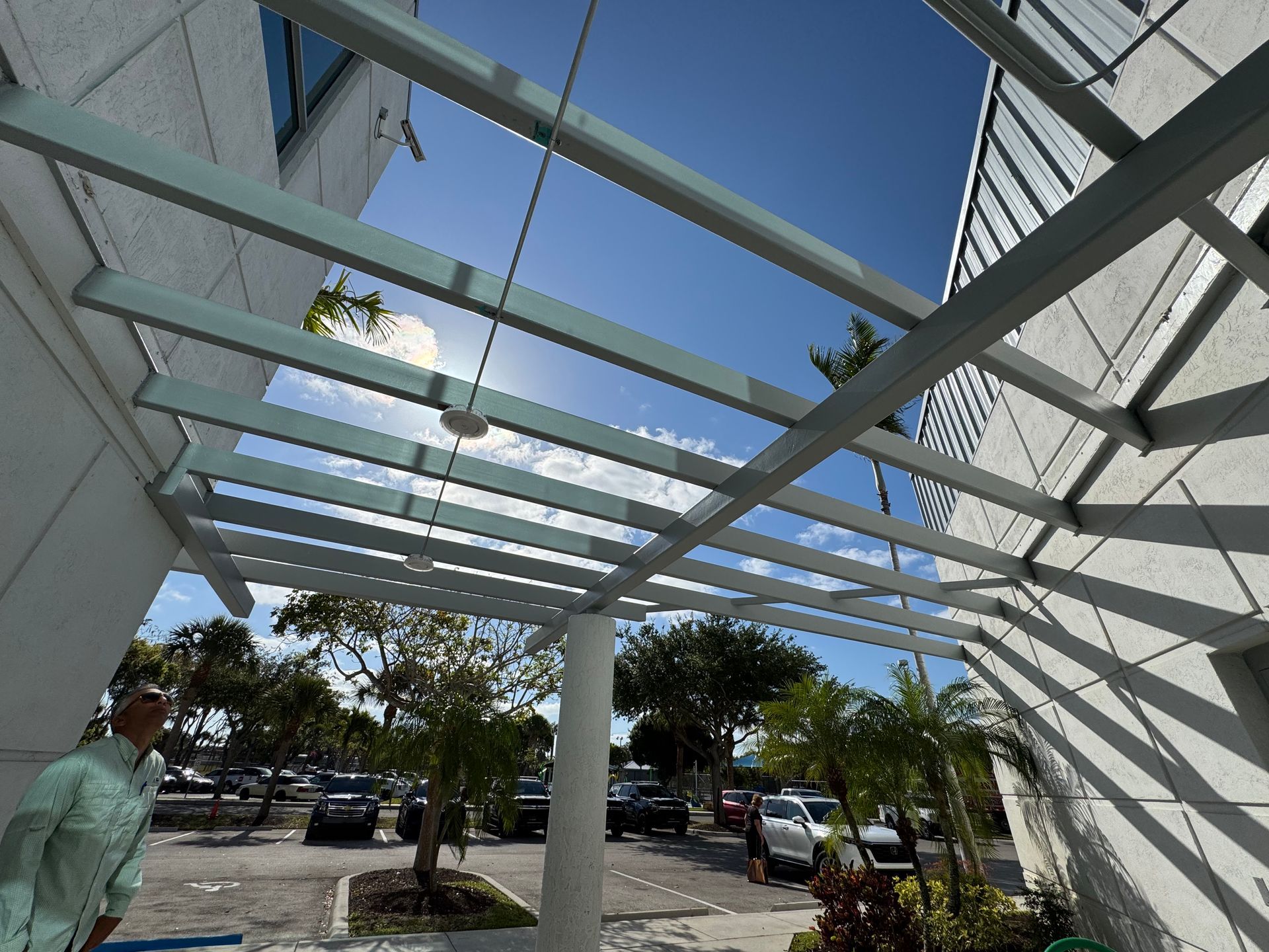 Pergola over building entrance. Bright sunlight, light-colored structure, man looking up, parking lot in background.