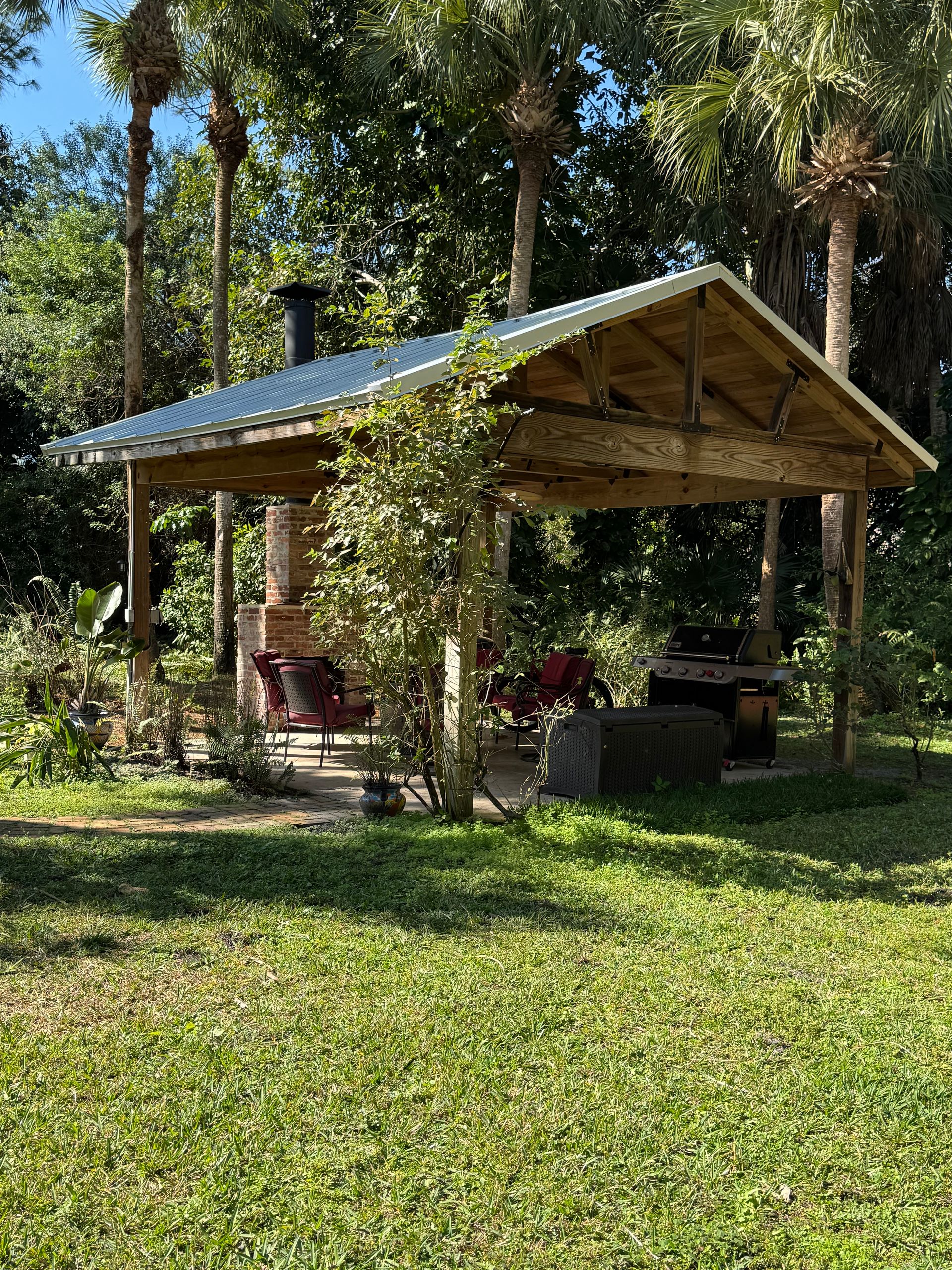 Wooden outdoor pavilion with a metal roof and a brick oven in a grassy area.