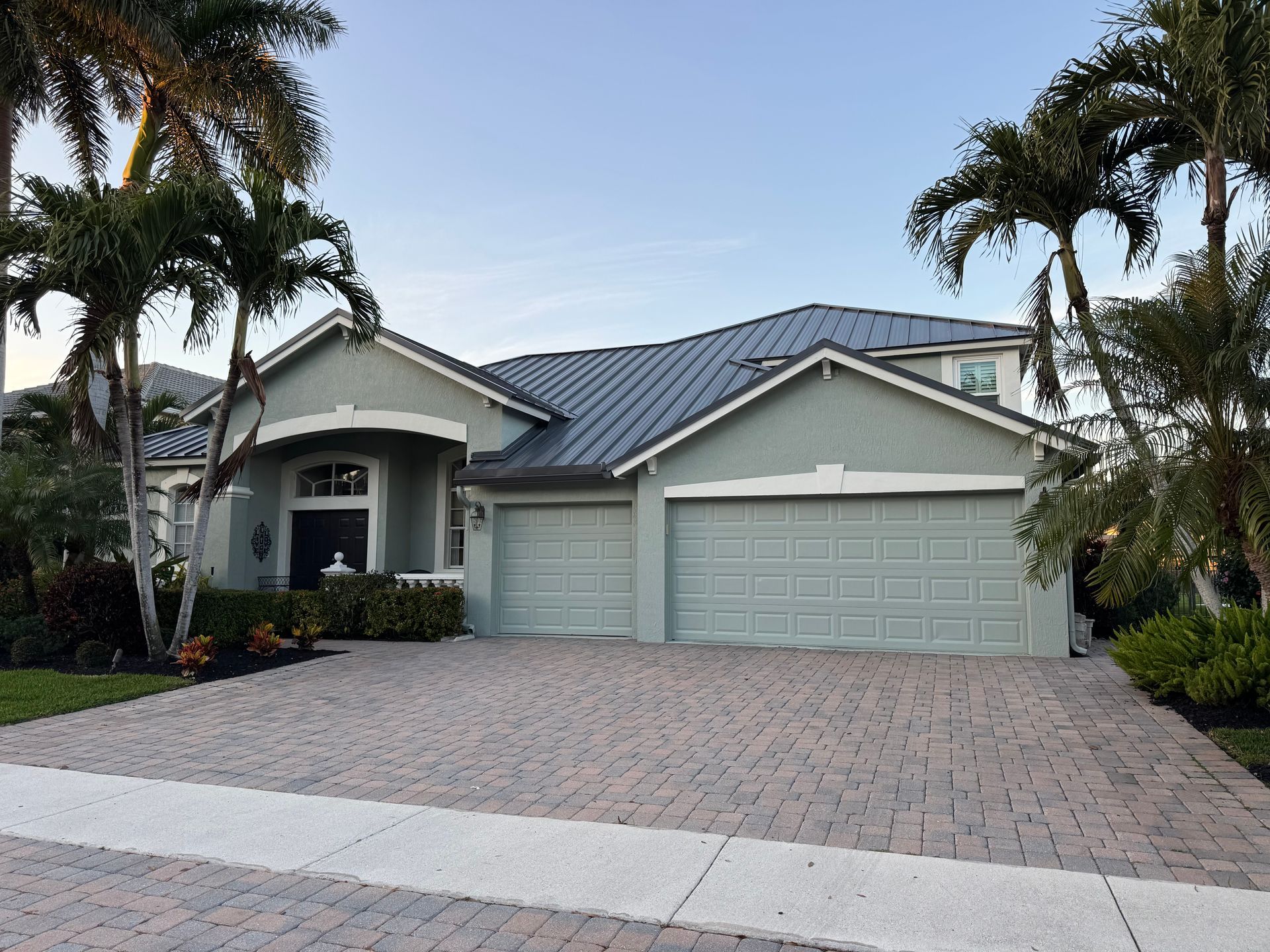 Two-story light green house with a brick driveway, palm trees, and a blue sky.