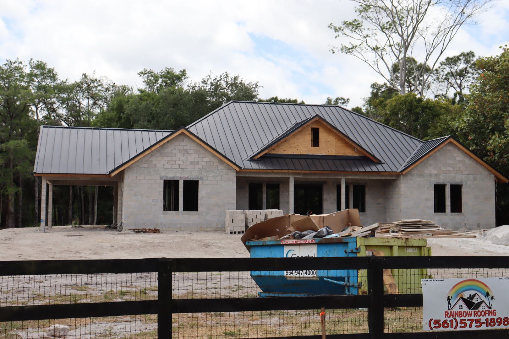 House under construction with metal roof, in a wooded area, blue dumpster in front.