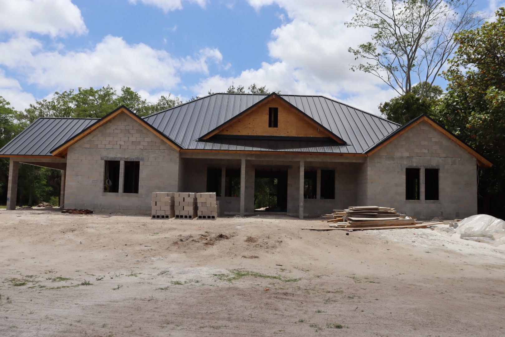 Unfinished house under construction with gray exterior, dark roof, and surrounding trees.