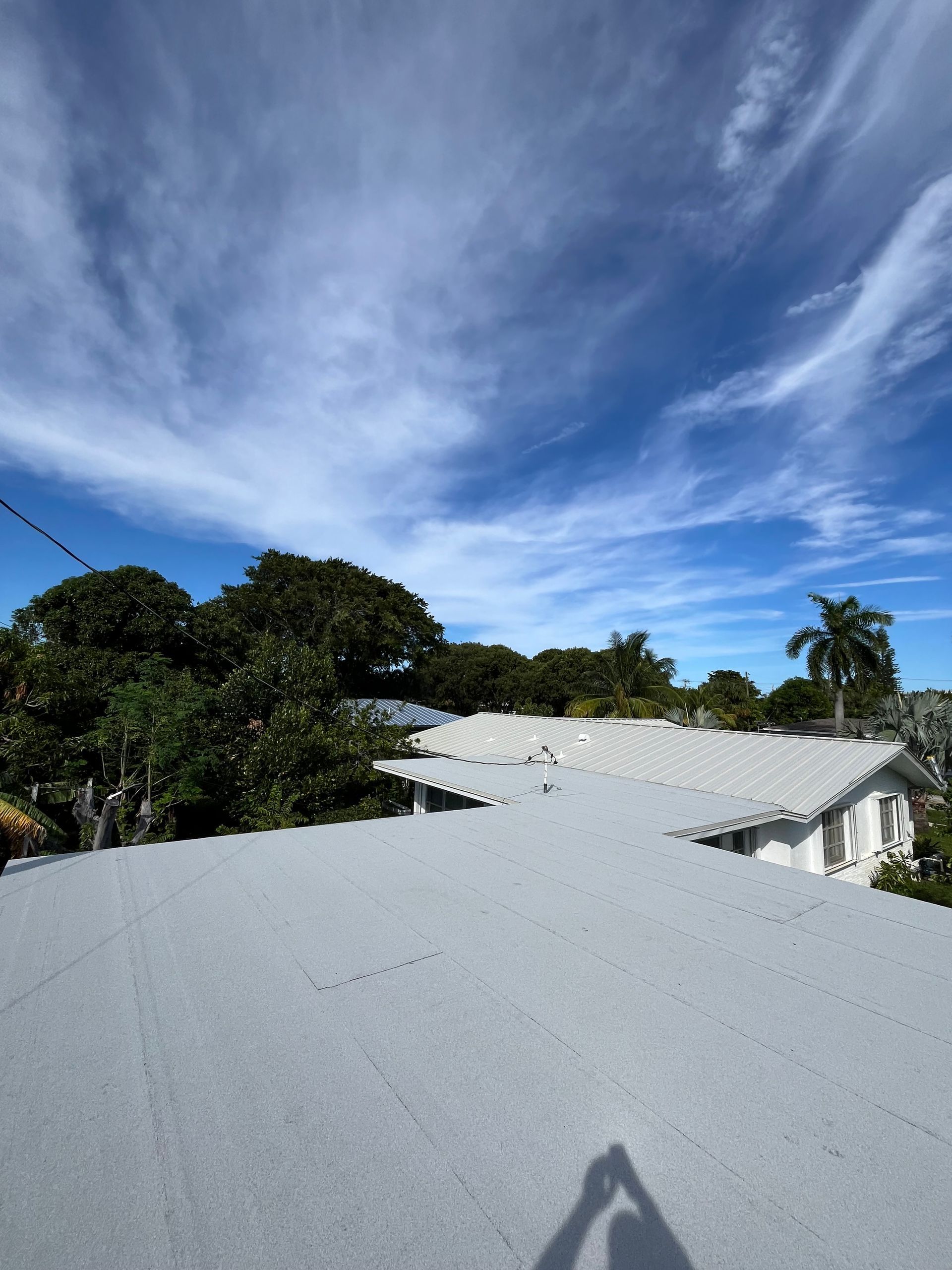 A gray roof under a blue sky with wispy clouds, trees and a house in the background.