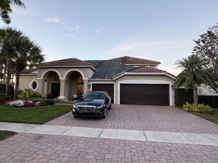 A luxury home with a black car parked in front on a brick driveway, palm trees.