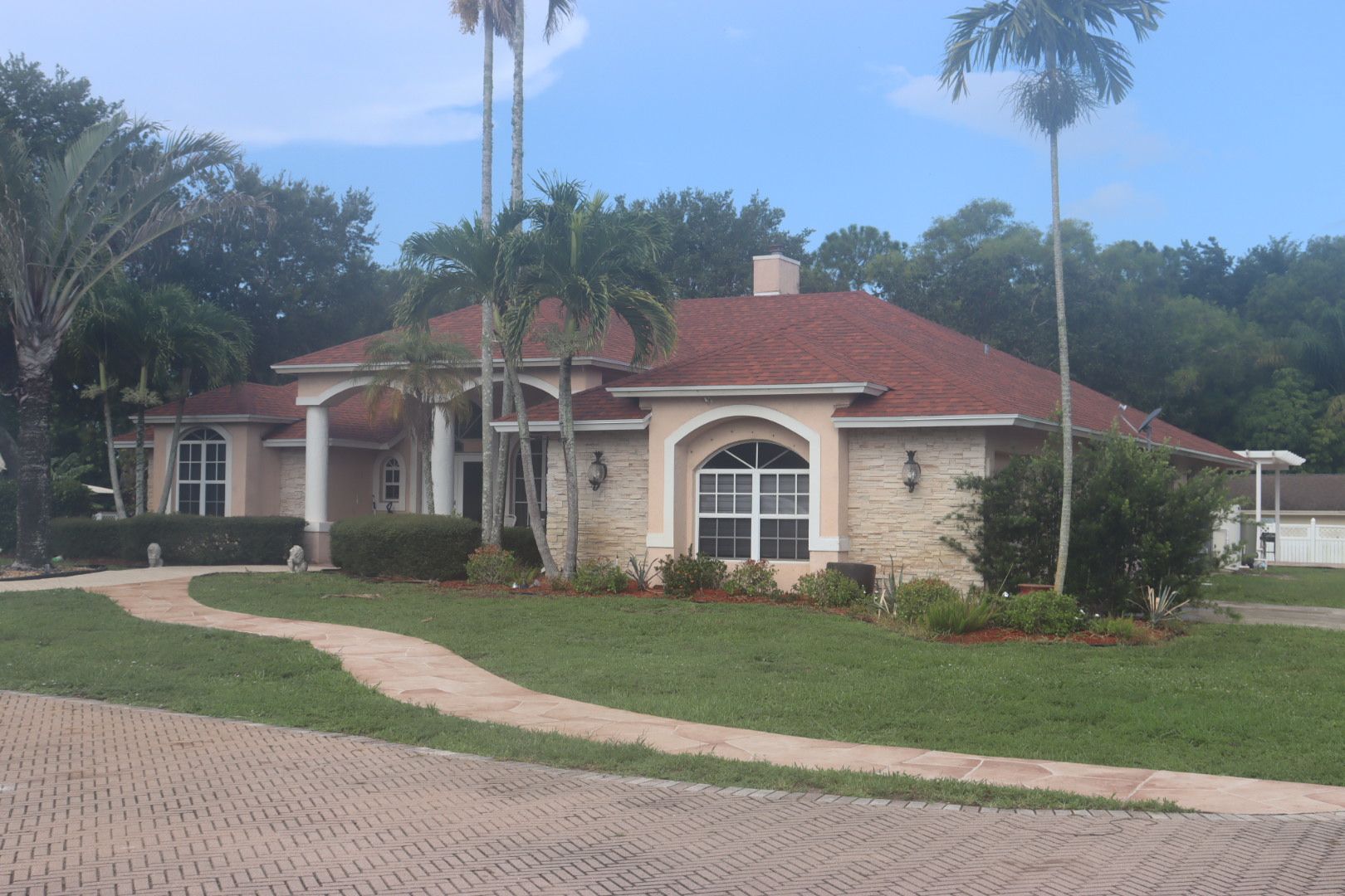 Tan house with red roof and palm trees, on a brick-paved driveway, set in a green, grassy yard.