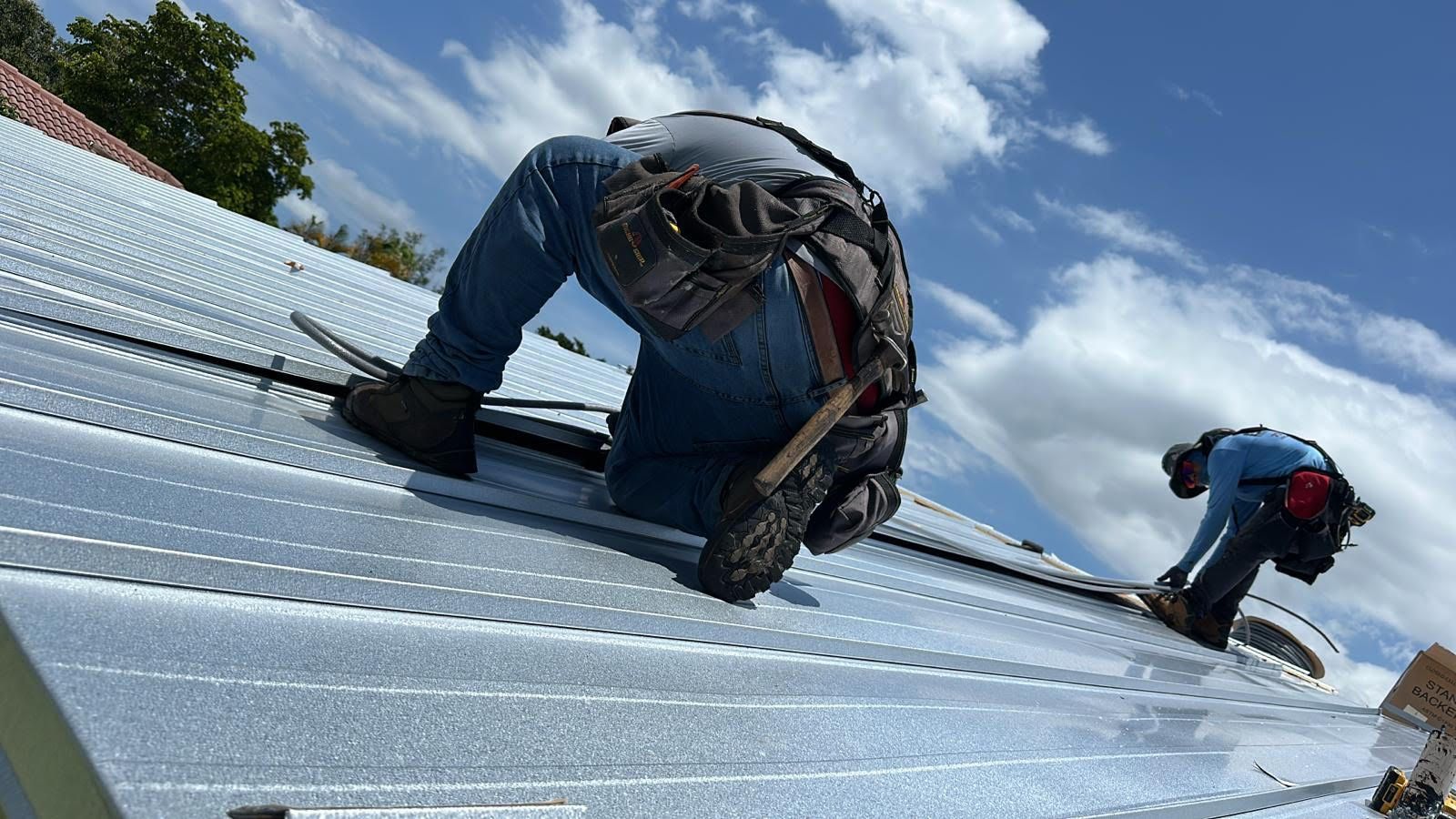 Two roofers installing metal panels on a rooftop under a partly cloudy sky.