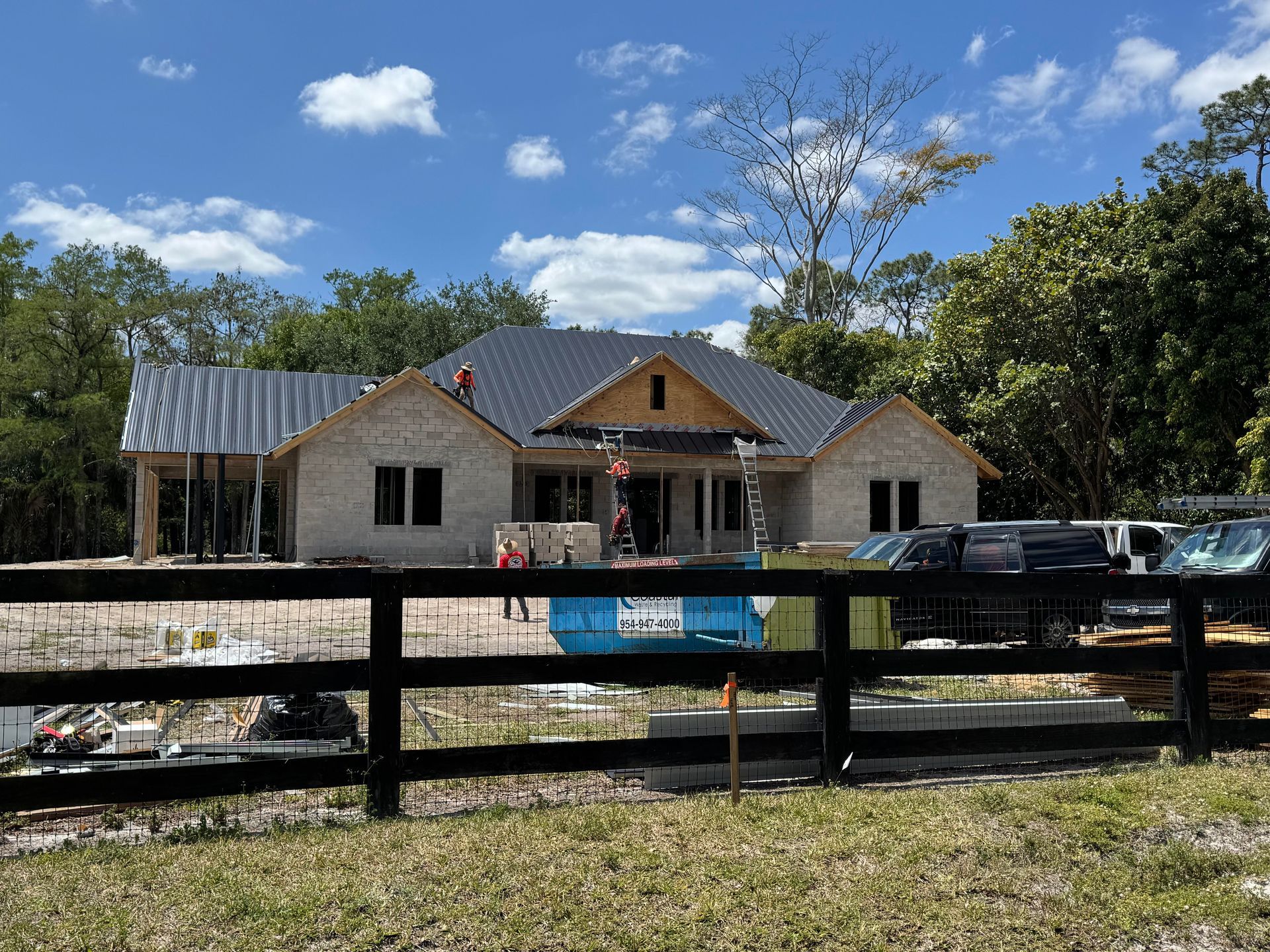 Construction site of a beige brick house with a dark gray roof, behind a black wooden fence, sunny day.