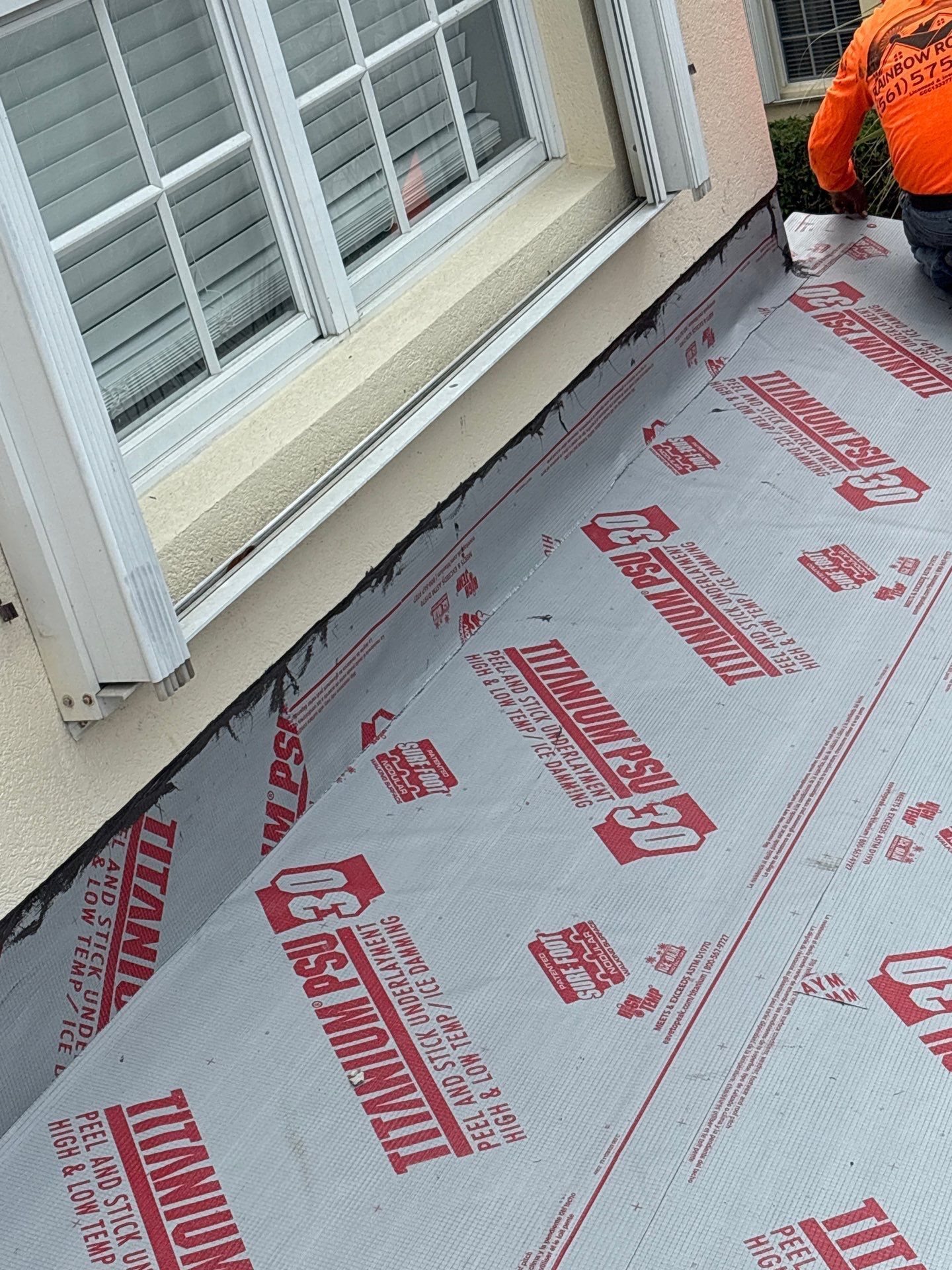 Roofers installing gray and red underlayment near a window.