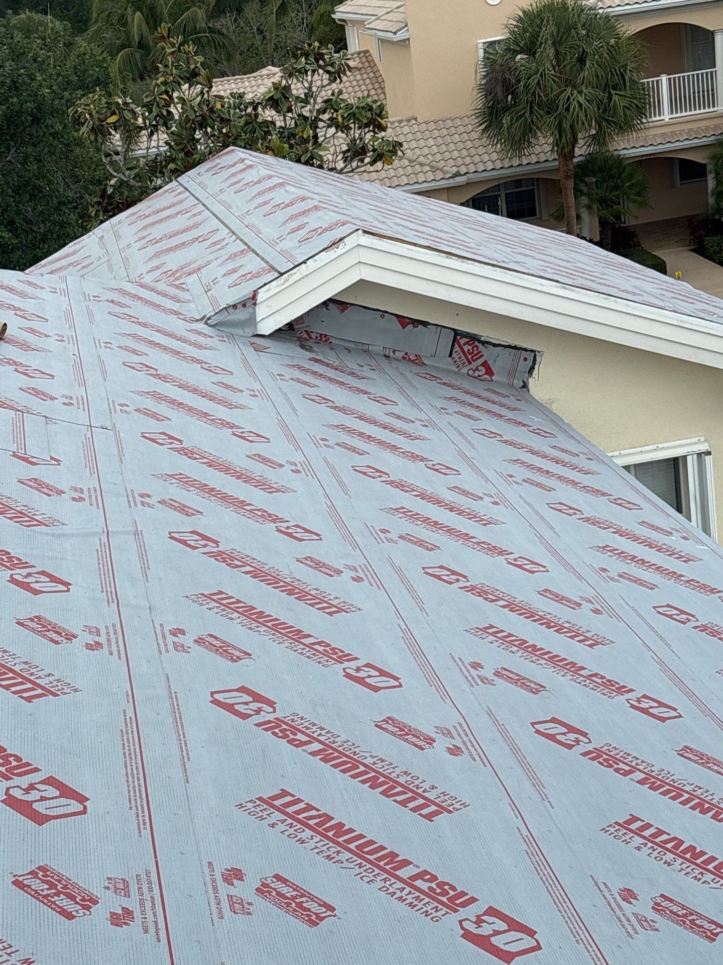 Roof covered with gray underlayment with red lettering, a white overhang, and a building in the background.