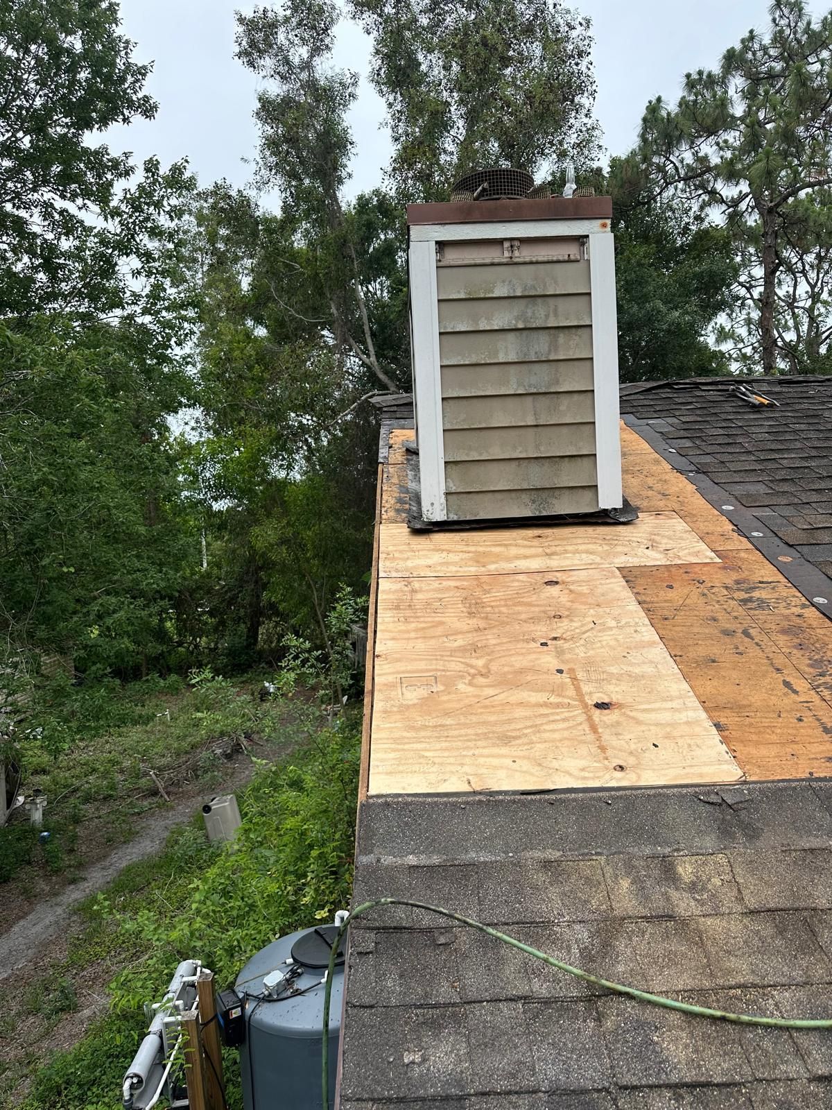 Chimney on a rooftop undergoing repairs. Shingles are damaged. Trees in the background.