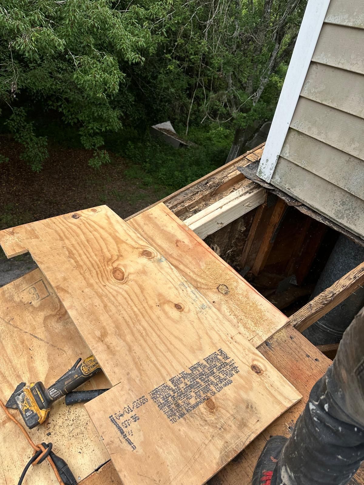 Rooftop repair: plywood over an opening, showing a toolbox and house siding in a green outdoor setting.