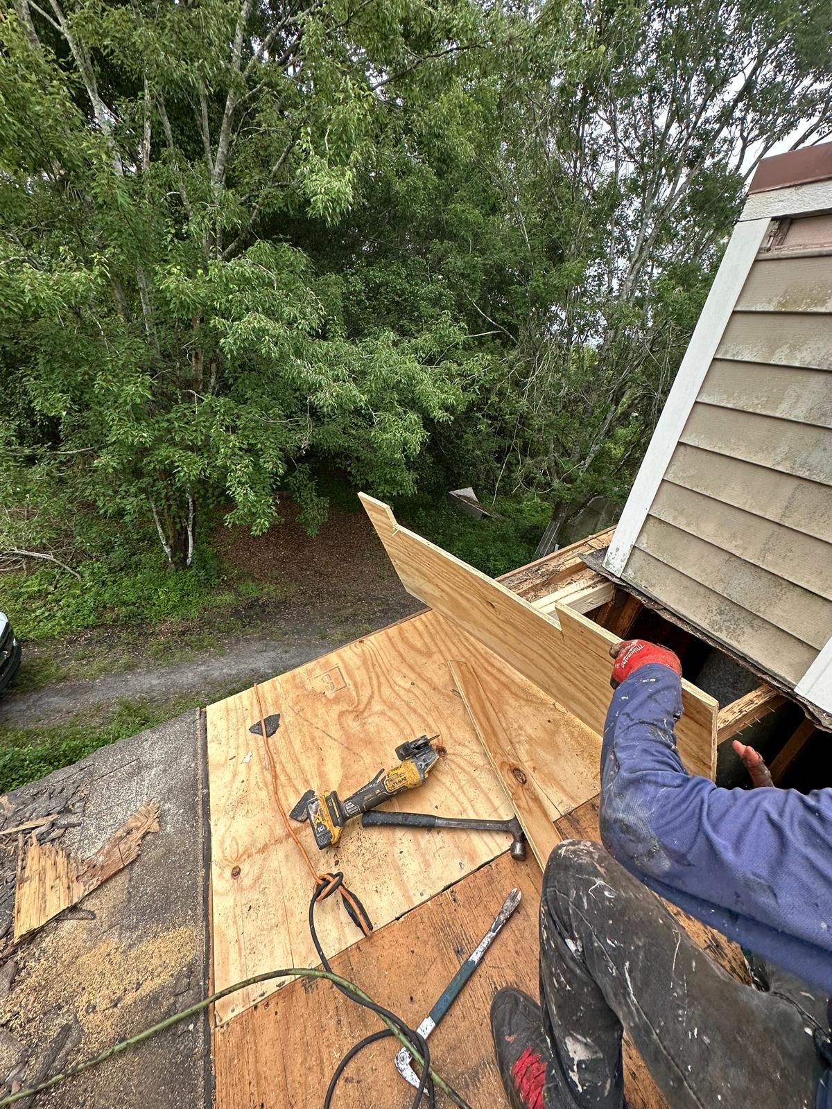 Roofer on a roof installing wood, with tools visible, trees in background.