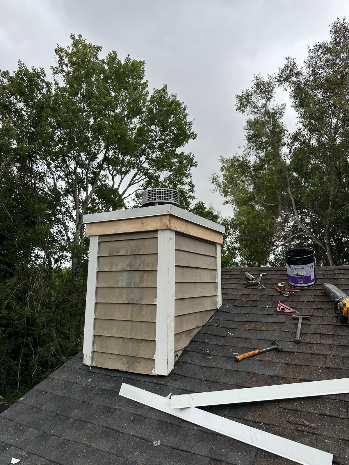 Chimney on a rooftop; clad in brown siding with white trim, trees in the background.