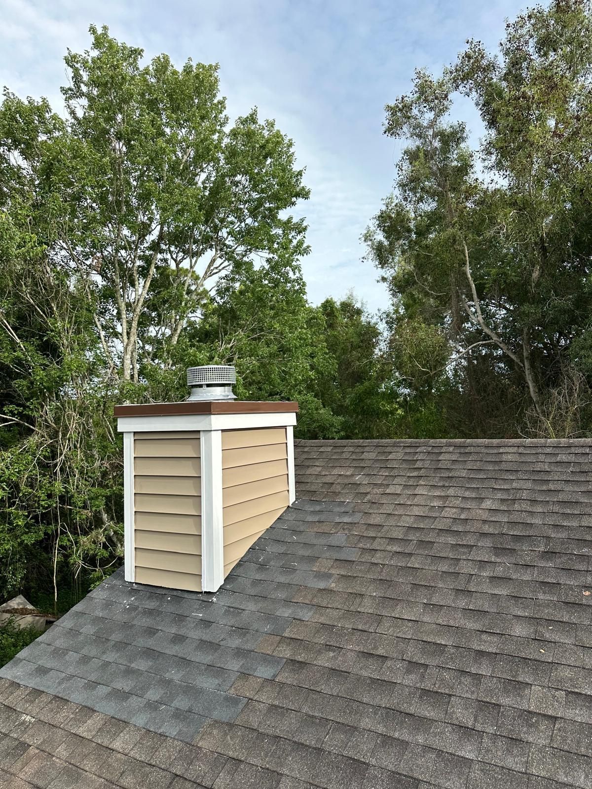 Chimney with light brown siding and a cap on a rooftop, against a backdrop of green trees and a cloudy sky.