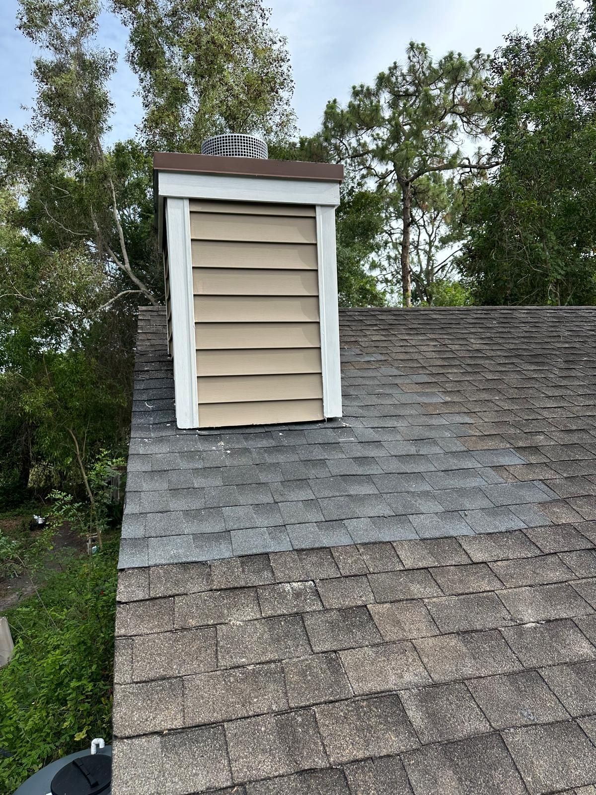 A weathered roof with shingles and a beige vent against a tree-filled backdrop.