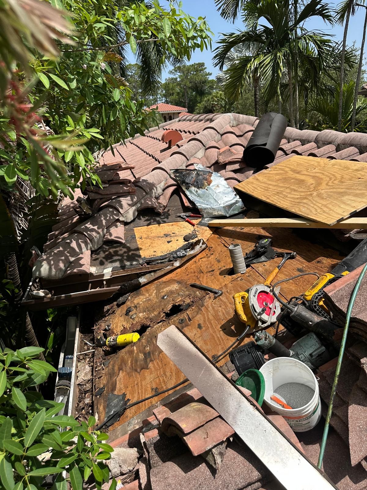 Roof damage, tiles removed, exposing rotted wood, surrounded by tools and debris, tropical setting.