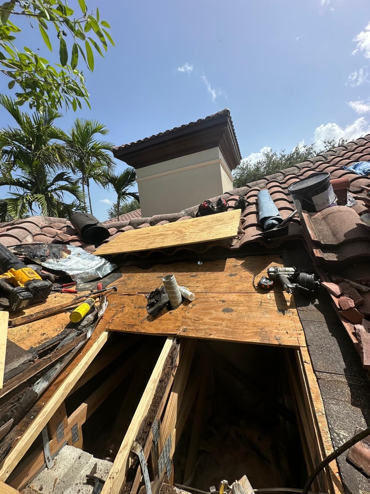 Rooftop with broken tiles, exposed wood, and tools near a chimney under a partly cloudy sky.