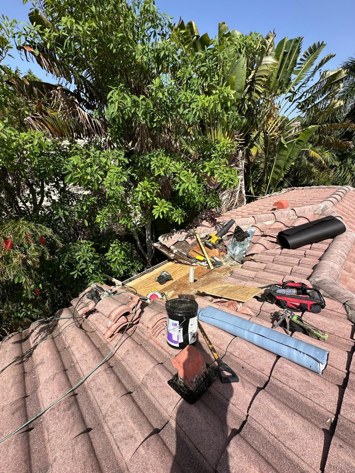 Roof with broken tiles, repair tools, and a container of black sealant. Green trees in the background.