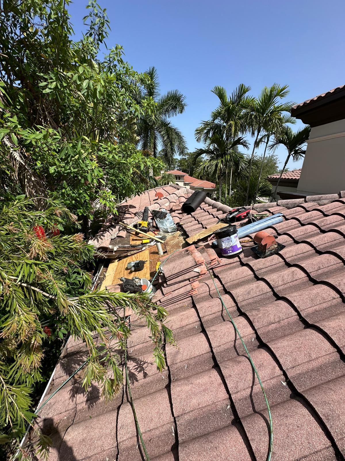 Roof damage with debris and tools, surrounded by greenery and palm trees under a blue sky.