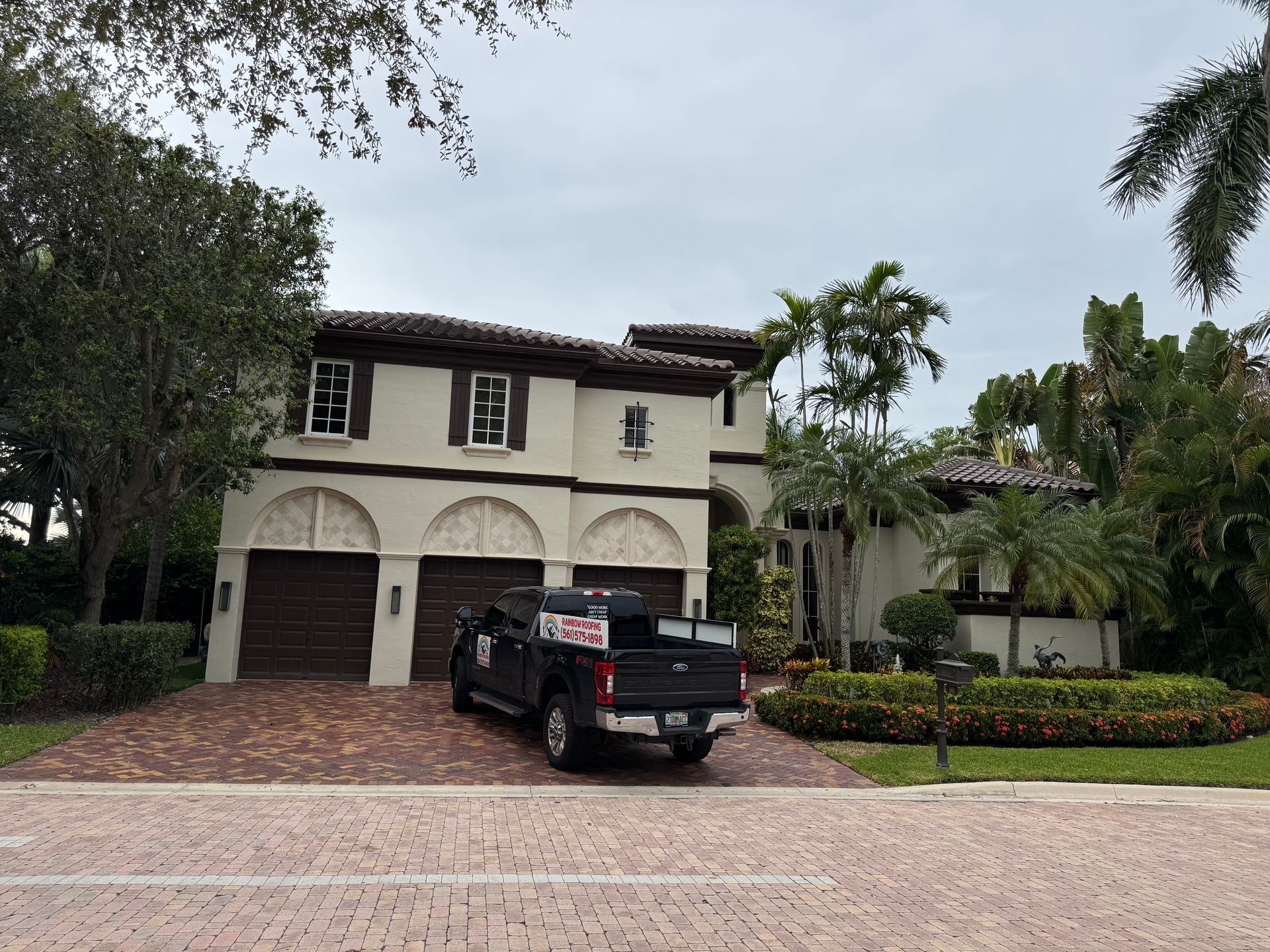 Two-story beige home with brown arched garage doors, palm trees, and a dark truck parked in front.