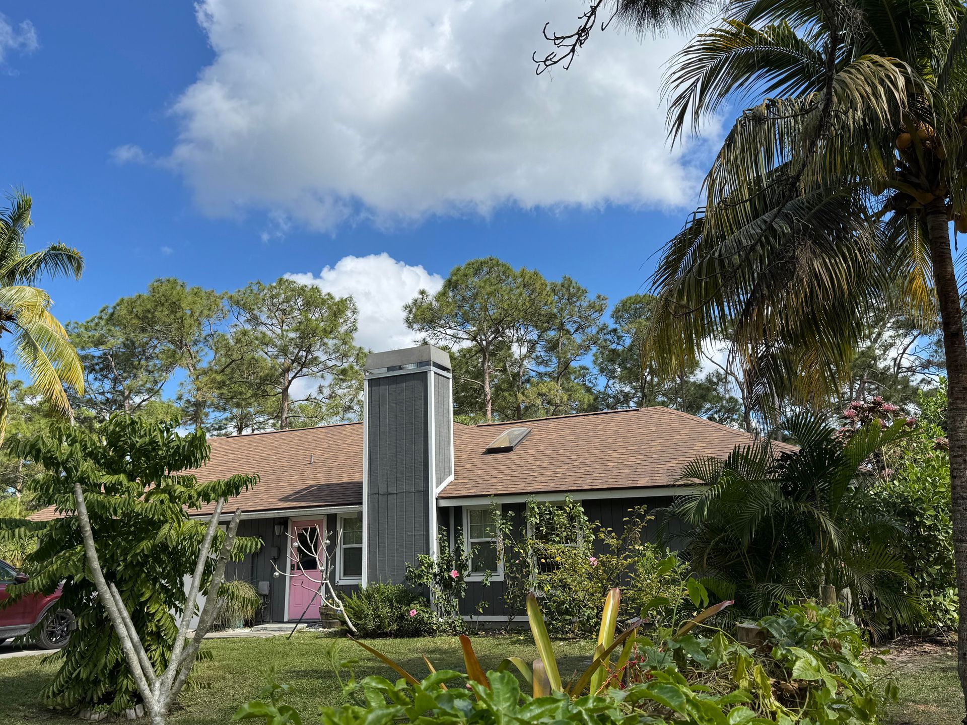 A gray house with a brown roof and tall chimney, surrounded by green trees and a blue sky with clouds.