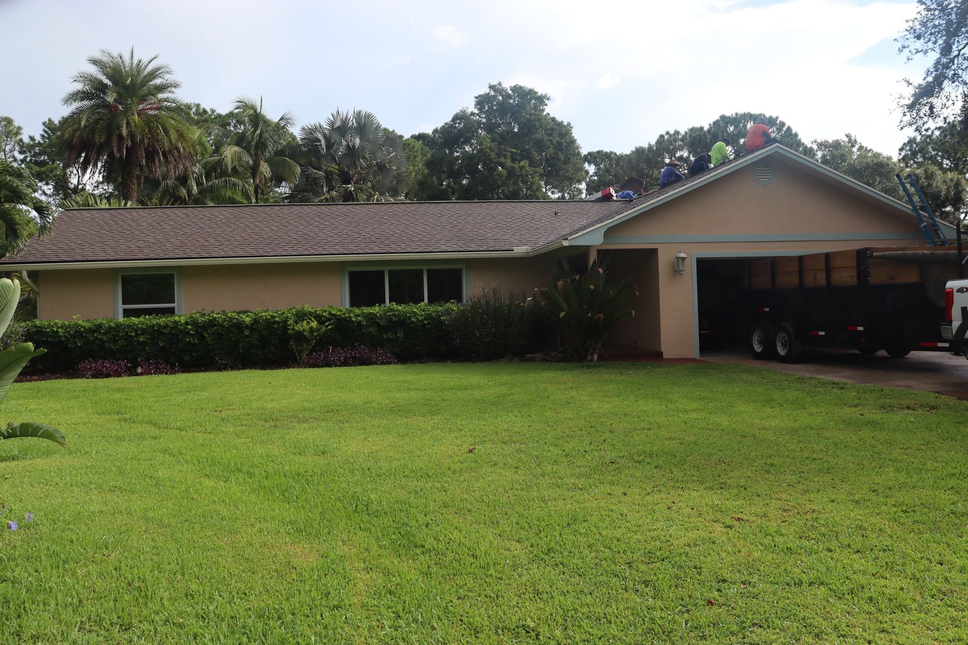 A roof being worked on by people; tan house, green lawn, dark roof. A trailer is parked beside the garage.