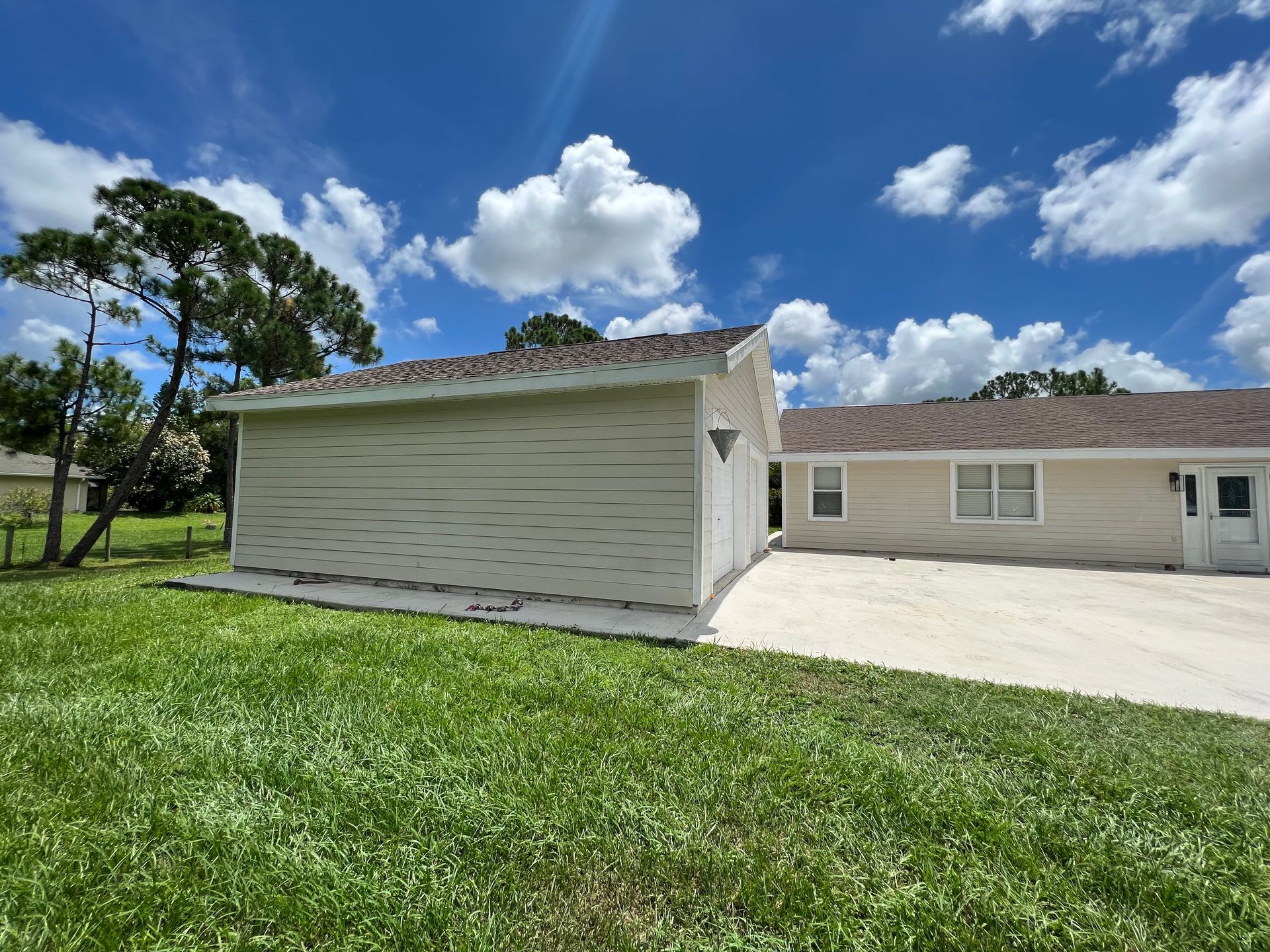 Tan building with a garage door, next to a house on a grassy lawn under a blue sky with clouds.