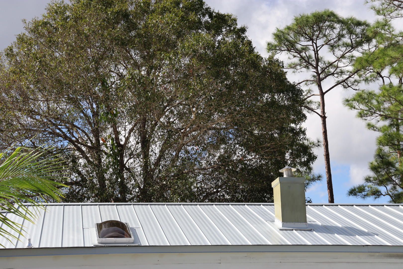 Metal roof with a chimney and skylight, with trees and a cloudy sky in the background.