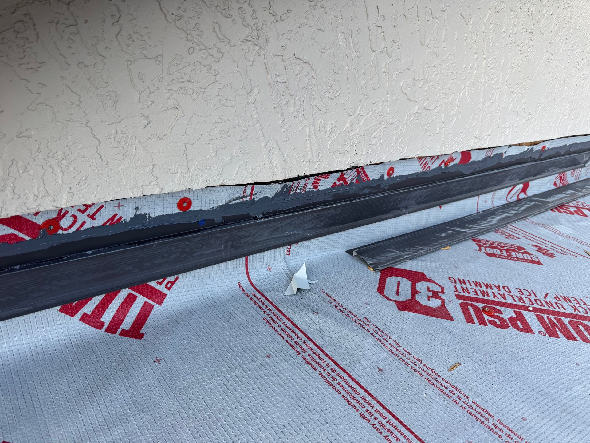 Close-up of a roof's edge with gray flashing and red-and-white underlayment next to a stucco wall.
