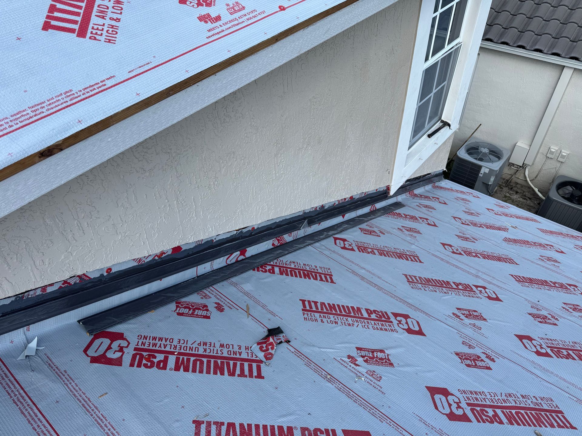 Roofing materials and flashing installed near a stucco wall and window. Red underlayment and dark flashing.