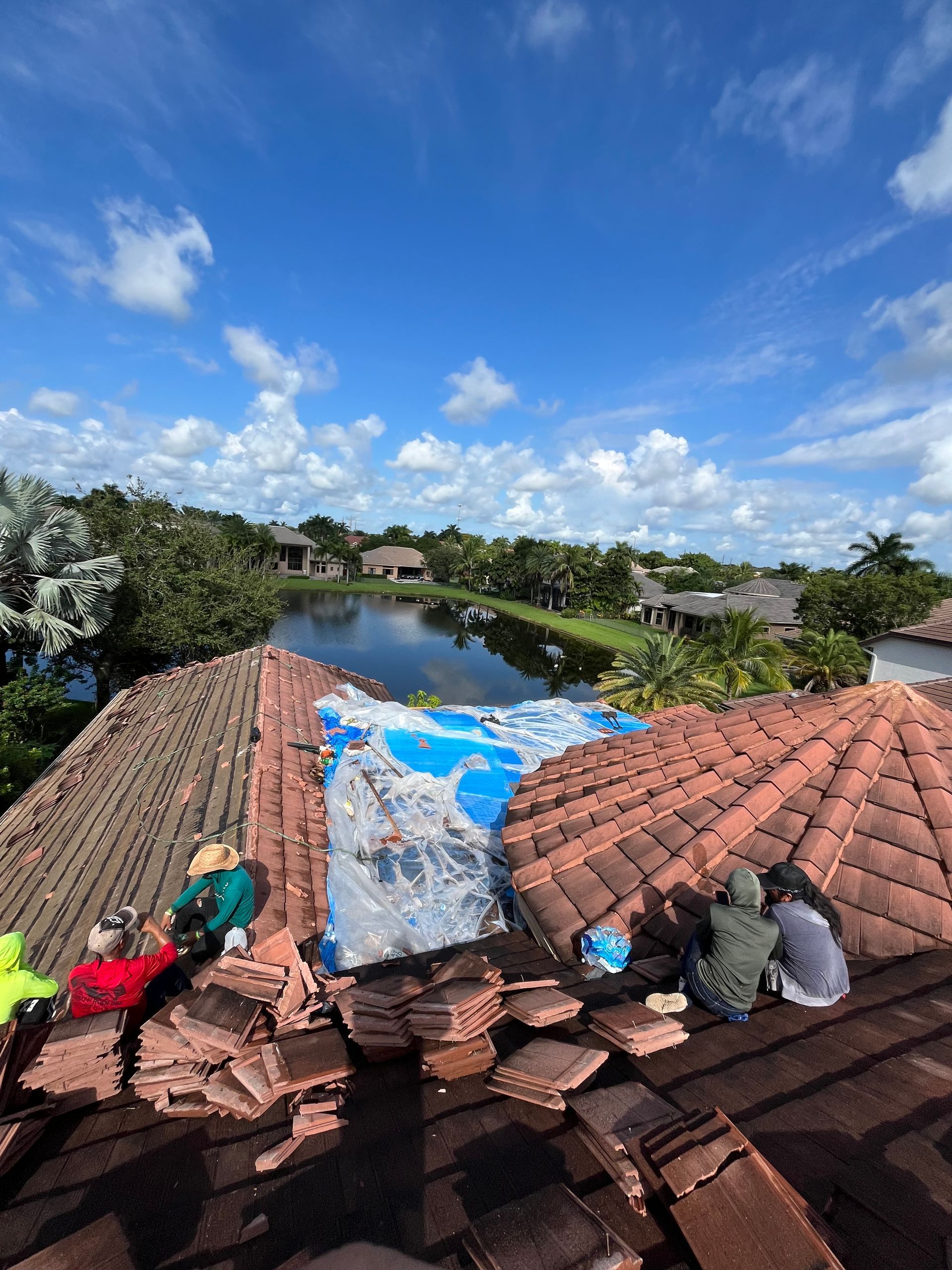 Workers on a clay tile roof, partially covered with tarp, overlooking a lake and houses on a sunny day.