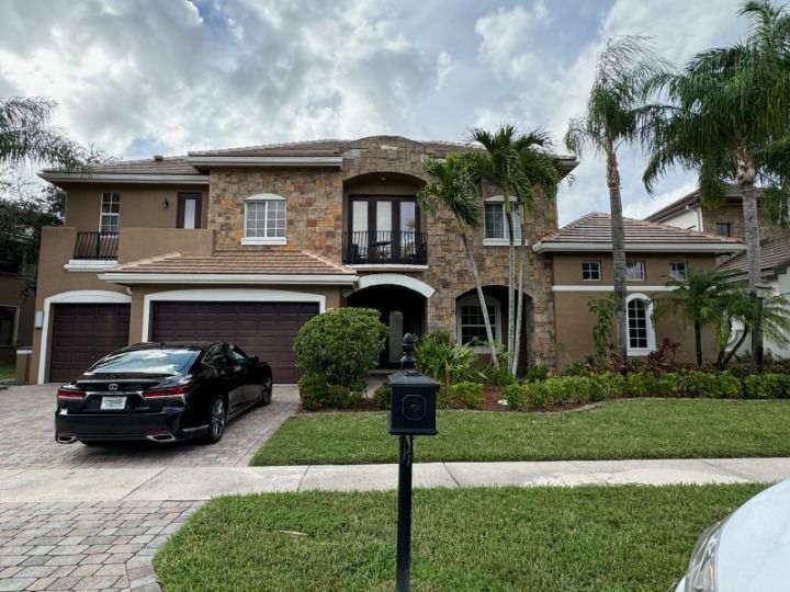 Luxurious two-story home with stone facade and a black car parked in front. Green grass and palm trees surround it.