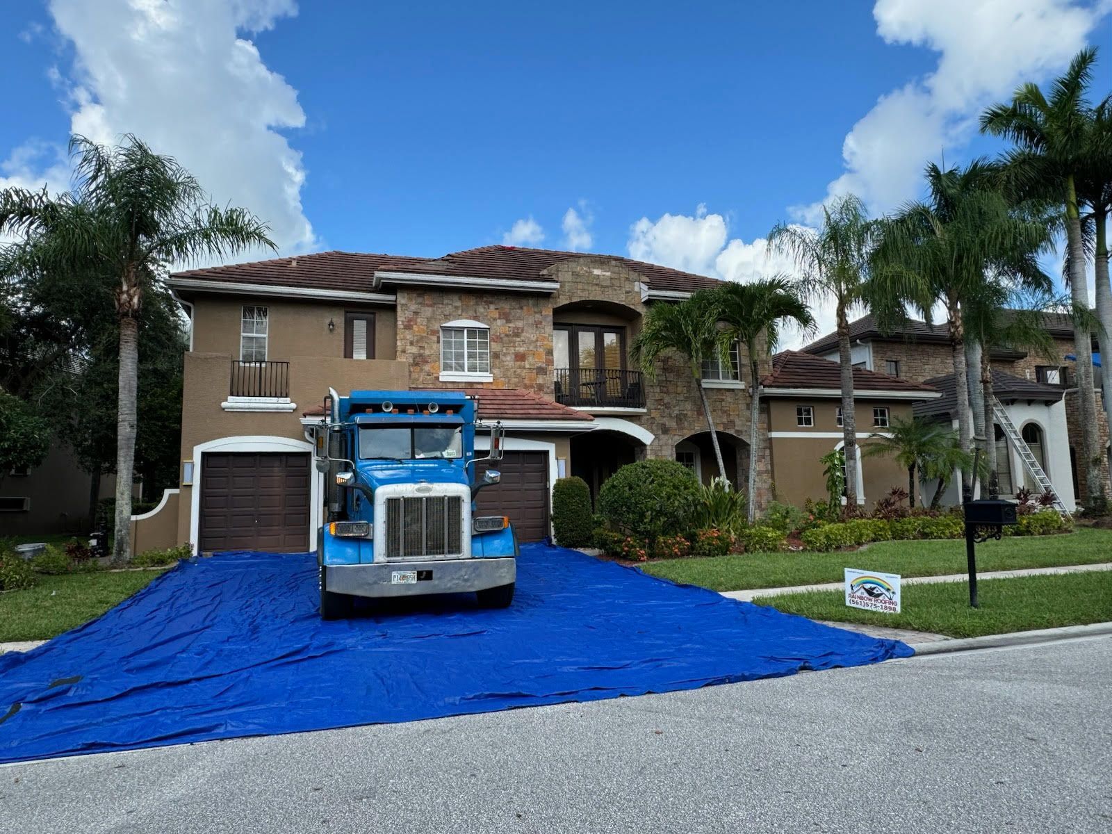 Blue truck parked on blue tarp in front of a two-story house with a stone facade and a brown roof.