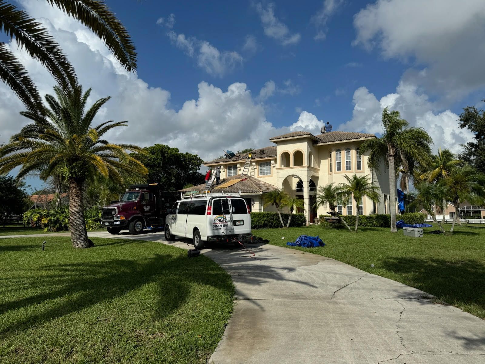 A large, yellow house with workers and vehicles in front, under a blue sky.