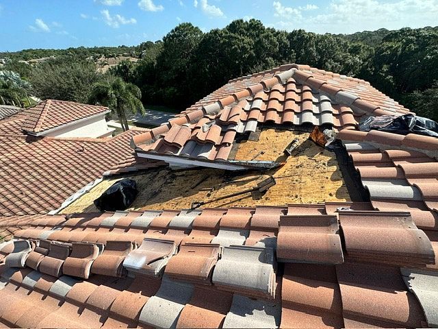 Roof damage: broken clay tiles on a partially exposed wooden roof, outdoors with blue sky and trees.