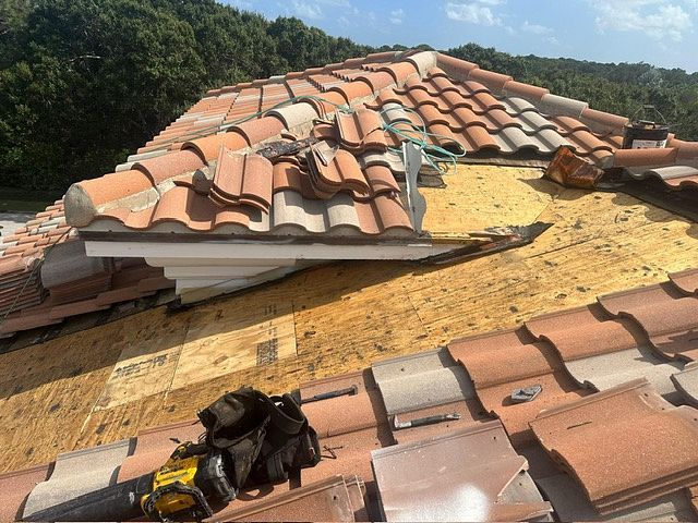 Roof with missing clay tiles, showing exposed plywood and tools. Orange tiles against blue sky.