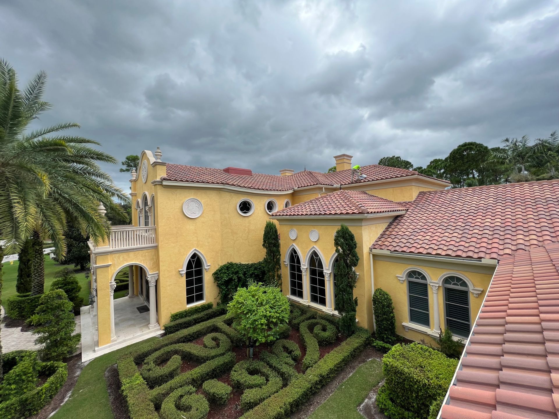 Yellow stucco mansion with red tile roof and ornate garden on a cloudy day.