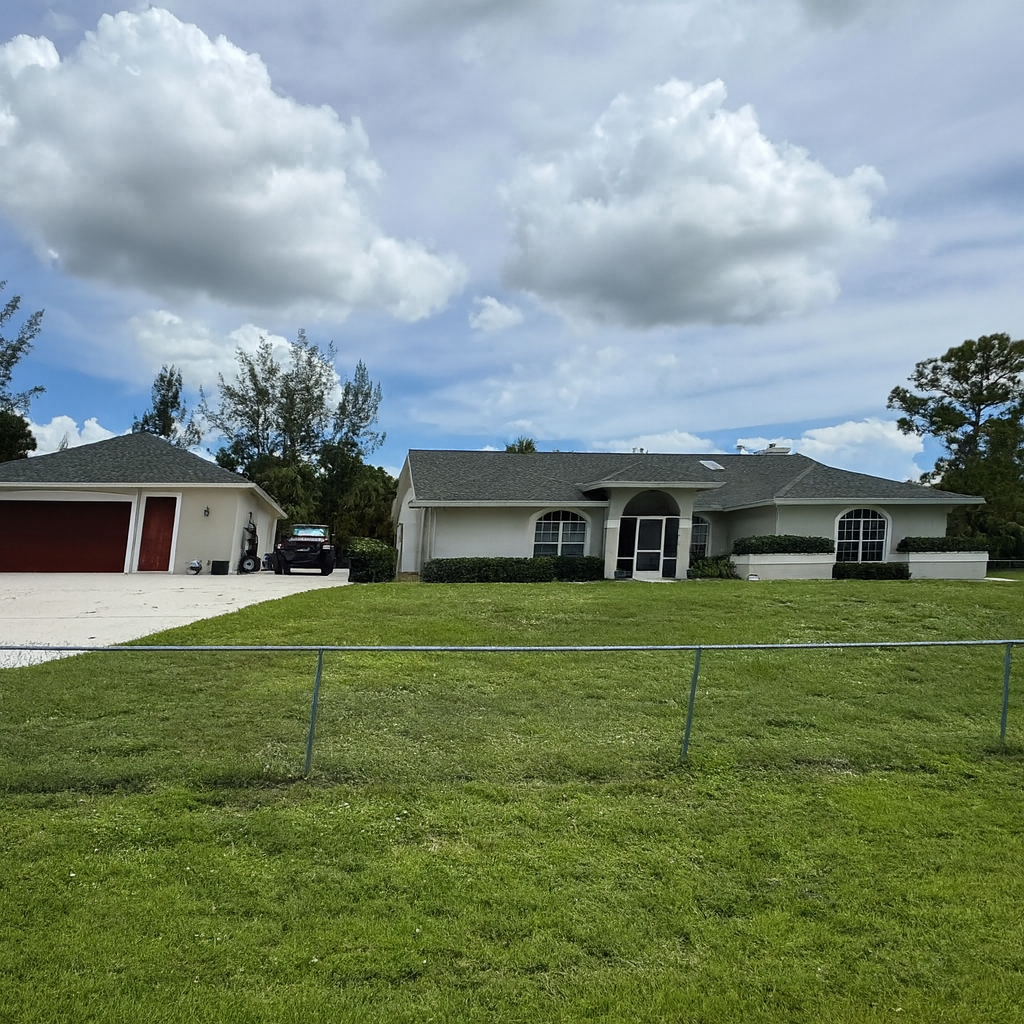 White house with a detached garage, green lawn, cloudy sky, chain-link fence.